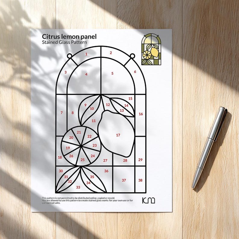 May include: A printed pattern for a citrus lemon stained glass panel. The design features a lemon and leaves within a window frame, with numbered sections for glass cutting. A silver pen rests on the wooden surface.