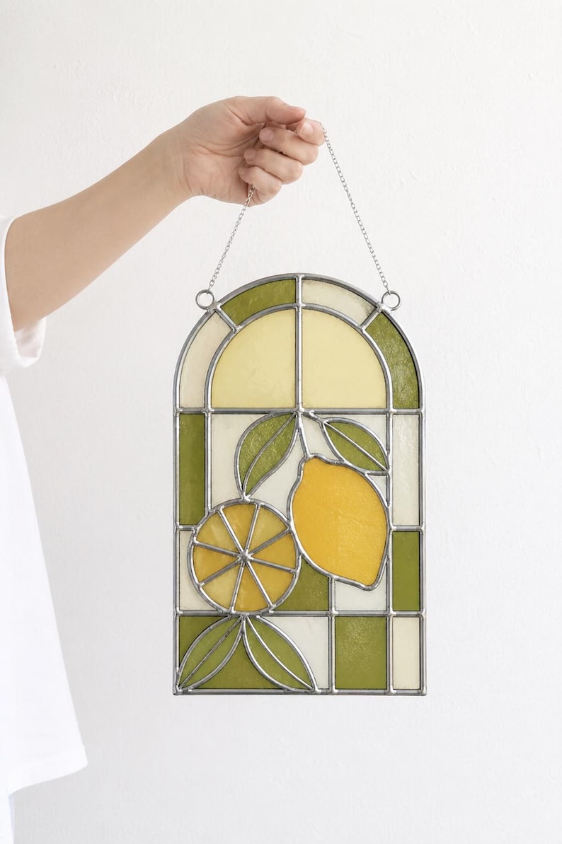 May include: Stained glass window panel featuring a lemon design. The panel has a rectangular shape with an arched top. The design includes a whole lemon, a lemon slice, and green leaves. The colors are yellow, green, and white.