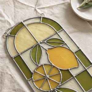 May include: Stained glass window panel featuring a lemon design. The panel has a silver metal frame and a chain for hanging. The design includes a whole lemon, a lemon slice, and green leaves, with yellow and green glass.