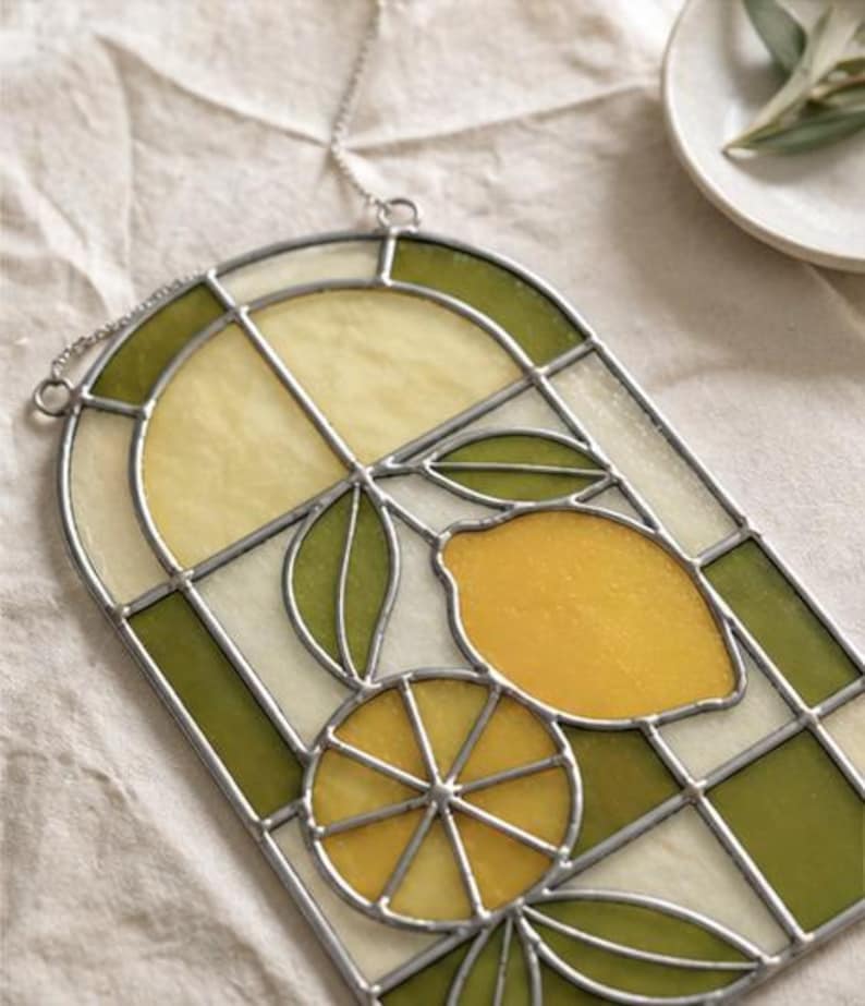 May include: Stained glass window panel featuring a lemon design. The panel has a silver metal frame and a chain for hanging. The design includes a whole lemon, a lemon slice, and green leaves, with yellow and green glass.