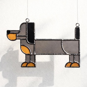 May include: A stained glass dachshund dog ornament. The dog is depicted in a geometric style with gray, black, and orange sections. It hangs from a thin wire, perfect for a window or wall decoration. The dog's body is approximately 8 inches long.