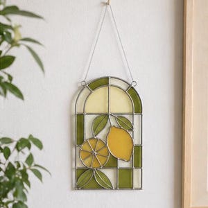 May include: Stained glass window panel featuring a lemon design. The panel has a silver metal frame with a green and yellow color scheme. The design includes two lemons and green leaves. The panel is hanging from a chain.