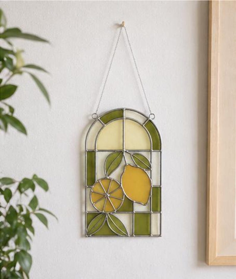 May include: Stained glass window panel featuring a lemon design. The panel has a silver metal frame with a green and yellow color scheme. The design includes two lemons and green leaves. The panel is hanging from a chain.