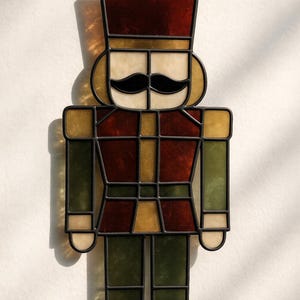 May include: Stained glass nutcracker figure with a black outline. The figure has a burgundy hat and jacket, with green and gold accents. The mustache is black, and the face is cream-coloured. A festive holiday decoration.