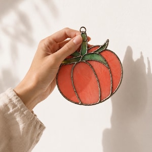 May include: A stained glass ornament shaped like a tomato with vibrant red glass segments and green glass leaves at the top. The pieces are joined by dark metal solder lines, and a small metal ring is attached to the top for hanging purposes.
