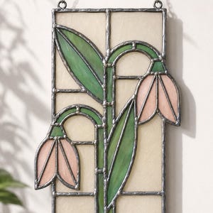 May include: A rectangular stained glass suncatcher featuring two drooping pink bell-shaped flowers with green stems and leaves, set against a cream-colored background. The glass pieces are joined by dark metal solder lines and hang from a metal chain.