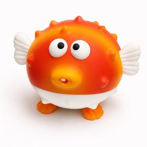 May include: A whimsical orange and yellow pufferfish toy with large, surprised eyes and white fins. The toy has a white diaper-like bottom and orange feet, creating a playful and cartoonish appearance. The toy is designed for children.