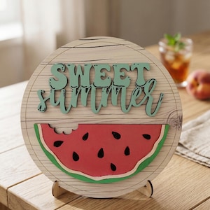 May include: A wooden round sign with the words "SWEET SUMMER" in green script. The sign features a watermelon slice with a bite taken out of it. The sign is propped up on a wooden stand and is on a wooden table.