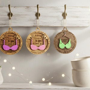 May include: Three wooden ornaments with bra designs hang from hooks. The ornaments are round with the text "A Good Friend Is Like A Good Bra." One bra is pink with white polka dots, one is solid pink, and one is green with white polka dots.