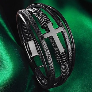 May include: A black leather bracelet featuring a silver cross design. The bracelet is made from multiple layers of leather and has a braided detail.