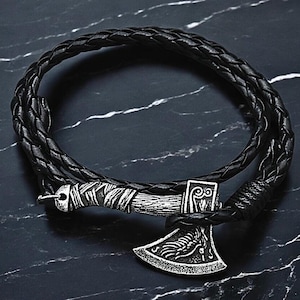 Puede incluir: Un brazalete trenzado de cuero negro con un dije de hacha de plata. El hacha tiene detalles intrincados y una envoltura de cuero trenzado en el mango.