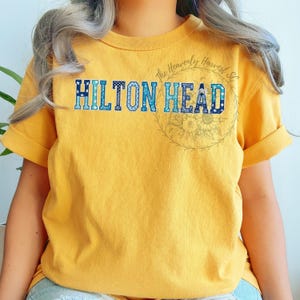 May include: Mustard yellow t-shirt with the text "HILTON HEAD" in blue, patterned lettering. The shirt has rolled-up sleeves and a floral design on the right side. The text "The Heavenly Harvest SC" is above the floral design.