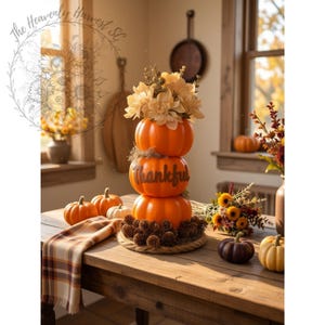 May include: A vertical stack of three bright orange pumpkins resting on a circular rope base with pinecones. The middle pumpkin features the word Thankful in dark script. The top pumpkin is topped with a bouquet of cream-colored flowers.
