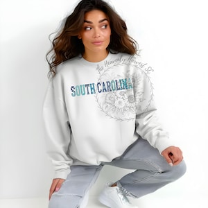 May include: White sweatshirt with the text "SOUTH CAROLINA" in blue and teal. The text is surrounded by a floral design. The sweatshirt is long-sleeved.