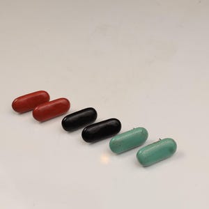 Può includere: Sei oggetti oblunghi a forma di pillola in fila. Due sono rossi, due neri e due verde acqua. Gli oggetti sono lisci e sembrano essere fatti di un materiale solido. Gli oggetti sono disposti diagonalmente su una superficie bianca.