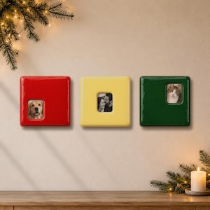May include: Three square ceramic photo frames in glossy red, yellow, and dark green. Each frame features a small, centered or offset rectangular cutout displaying a photograph: a golden retriever, a couple, and a cat, respectively.