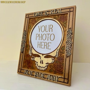 May include: Wooden photo frame with a Steal Your Face skull design. The frame features the text "LOVE IS REAL" and "NOT FADE AWAY". The center has a white circle with the text "YOUR PHOTO HERE". Decorative roses are carved into the frame.