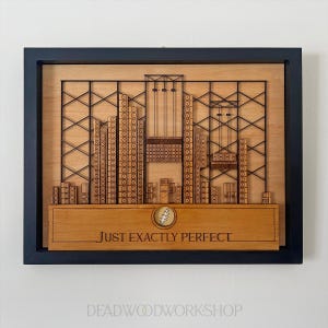 May include: Wooden wall art featuring a laser-cut cityscape design with a black frame. The artwork includes the text "JUST EXACTLY PERFECT" and a symbol. The piece is made of light brown wood.