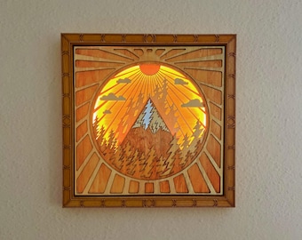 Può includere: Un'opera d'arte murale in legno con una scena di montagna e un sole con raggi. L'opera è incorniciata in una cornice di legno marrone chiaro.