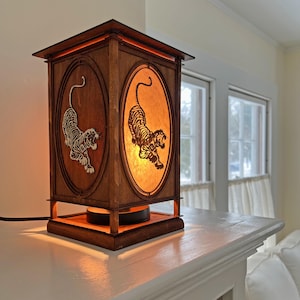 May include: A wooden lamp with a square base and a rectangular shade. The shade has two cut-out images of tigers on each side. The lamp is lit and casting a warm glow.