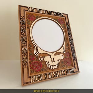 May include: Wooden picture frame with a skull design and the text "MAY THE FOUR WINDS BLOW YOU SAFELY HOME." The frame features a central circular opening, red roses, and floral accents. The frame is made of light brown wood.
