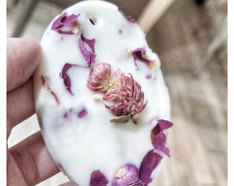 May include: A white oval-shaped wax melt with dried rose petals and a small pink flower embedded in the wax. The wax melt is being held in a hand.