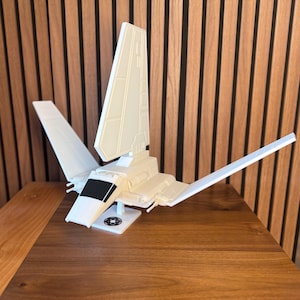 May include: A white model of a space shuttle with angled wings and a central fin. The model is on a stand with a circular emblem. The shuttle is set against a wooden background.