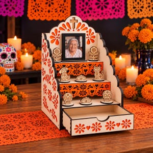 May include: A tiered wooden altar display featuring laser-cut floral patterns, a central photo frame, and small decorative figures of a dog, cat, and skulls. The structure includes three shelves and a bottom drawer with a cutout handle.