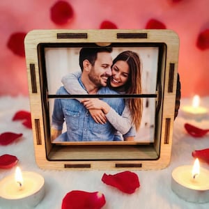 Può includere: Una cornice in legno con una foto di una coppia sorridente, circondata da petali di rosa e candele accese. La cornice ha un meccanismo rotante. Lo sfondo è rosa tenue.