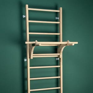 Könnte beinhalten: Holz-Sprossenwand mit Klimmzugstangenaufsatz, an einer grünen Wand montiert. Das Klettergerüst hat mehrere horizontale Sprossen zum Klettern und Trainieren. Die Klimmzugstange ragt nach außen und bietet zusätzliche Trainingsmöglichkeiten.
