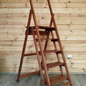 May include: A wooden step ladder with a dark brown finish. The ladder has four steps and a top rail, and is designed to fold for storage. The ladder is made of wood and has a classic design.