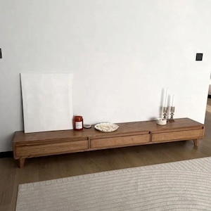 May include: A long, low, wooden media console with three drawers and tapered legs. A small red jar, a decorative plate, and a candle holder with three candles sit on top. A white canvas and a rug are in the background.