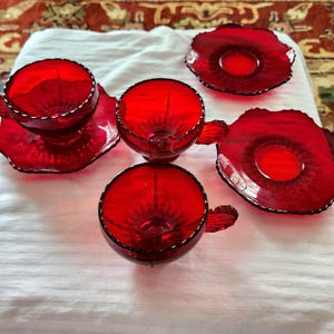 May include: A set of three translucent ruby red glass teacups with matching handles and two matching saucers. Each piece features a scalloped edge and a pressed glass pattern radiating from the center, creating a vibrant, deep red display.