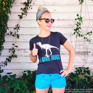 May include: A dark gray t-shirt with a white dinosaur graphic and the text "I GIVE HUGS" in blue. The person is wearing blue shorts, sunglasses, and a gray headband. Photo courtesy of The Band Wife, T-shirt design by Skip N Whistle.