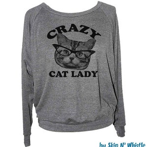 May include: Gray long-sleeved sweatshirt with the words "CRAZY CAT LADY" above and below a cat wearing glasses. The sweatshirt has a scoop neck and fitted cuffs. The design is in black.