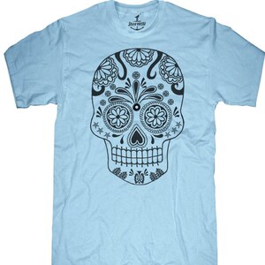 May include: A light blue t-shirt featuring a detailed black sugar skull design. The skull has floral and geometric patterns, including flower-shaped eyes and a heart-shaped nose. The shirt has a crew neck and short sleeves. The brand name "SKULLHOUSE" is printed on the collar.