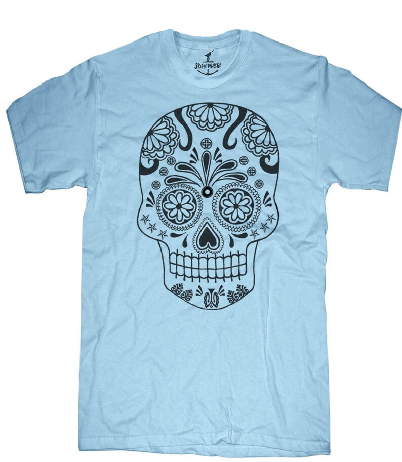 May include: A light blue t-shirt featuring a detailed black sugar skull design. The skull has floral and geometric patterns, including flower-shaped eyes and a heart-shaped nose. The shirt has a crew neck and short sleeves. The brand name "SKULLHOUSE" is printed on the collar.
