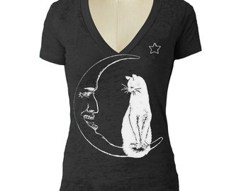 Puede incluir: Camiseta negra con cuello en V con un gráfico blanco de un gato sentado en una luna creciente con una cara. Una estrella blanca está a la derecha del escote. La camiseta se muestra en un maniquí.