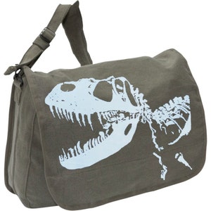 May include: Olive green messenger bag with a light blue Tyrannosaurus Rex skeleton graphic. The bag has an adjustable shoulder strap and a flap closure. The bag is made of a durable canvas material.