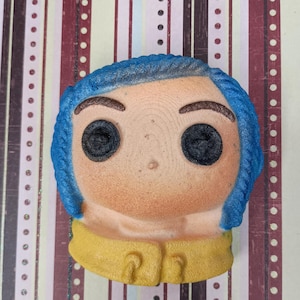 May include: A blue and yellow bath bomb shaped like a cartoon character with big eyes and a frown. The character has blue hair and is wearing a yellow shirt.
