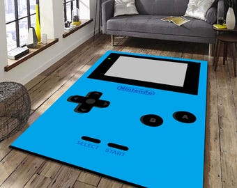 May include: A blue and black Game Boy-style rug with the word "Nintendo" printed on it. The rug features a directional pad, buttons labeled "A" and "B", and buttons labeled "Select" and "Start".