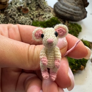 May include: A small, crocheted mouse figure. The mouse is cream-coloured with pink ears, paws, and nose. It has black eyes and a long, pink tail. The tiny mouse is held in a person's hand, with a blurred background of moss and wood.