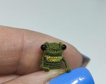 Op de afbeelding: Een kleine, met de hand gehaakte kikker in groen en geel. Het miniatuuramfibie heeft zwarte ogen en zit op een oppervlak. Dit unieke item is een charmant voorbeeld van handgemaakte kunst.