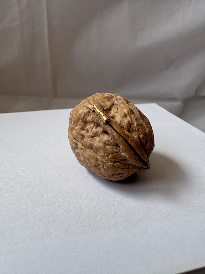 Open miniature walnut shell box with a small brass hinge, placed on a soft white fabric background. The natural wood texture and warm brown color of the walnut shell are clearly visible. A tiny handmade keepsake box perfect for storing rings or tiny treasures.