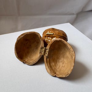 Open miniature walnut shell box with a small brass hinge, placed on a soft white fabric background. The natural wood texture and warm brown color of the walnut shell are clearly visible. A tiny handmade keepsake box perfect for storing rings or tiny treasures.