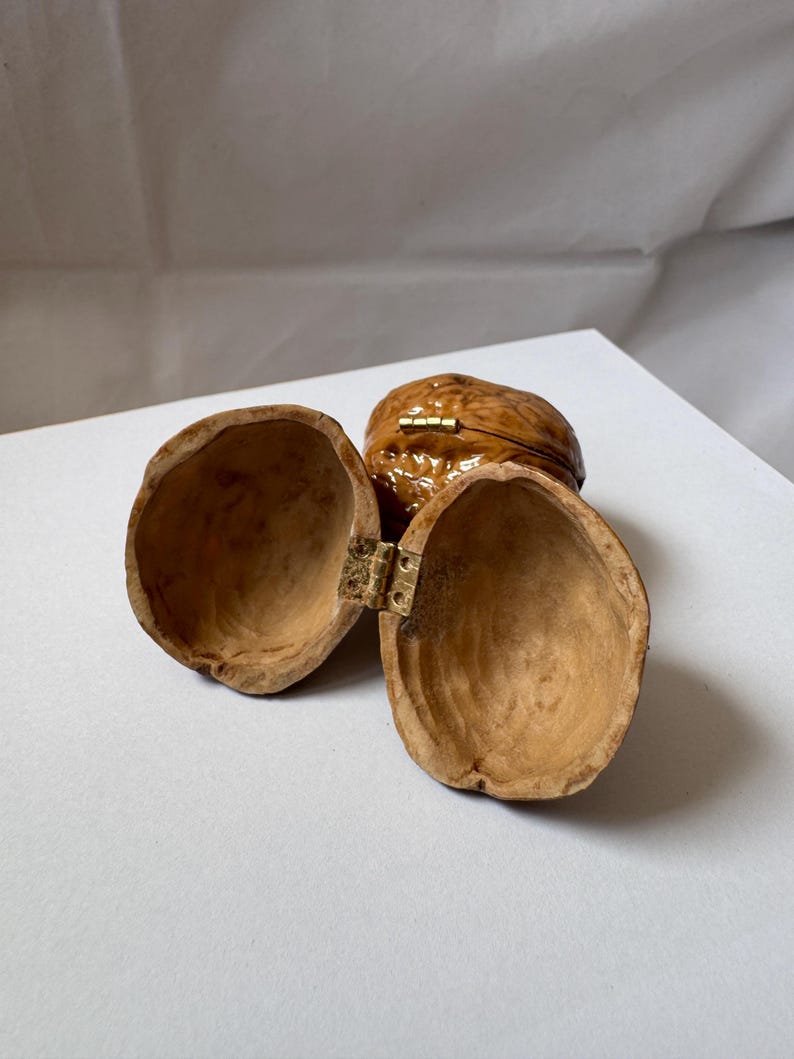 Open miniature walnut shell box with a small brass hinge, placed on a soft white fabric background. The natural wood texture and warm brown color of the walnut shell are clearly visible. A tiny handmade keepsake box perfect for storing rings or tiny treasures.
