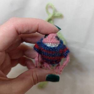 May include: A small crocheted creature with a pink body, blue and red striped sweater, and a toothy grin.