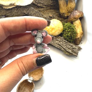 May include: A miniature crocheted gray mouse with pink ears and a pink tail. The mouse is held near a walnut shell, mushrooms, and moss. The background includes bark and other natural elements.