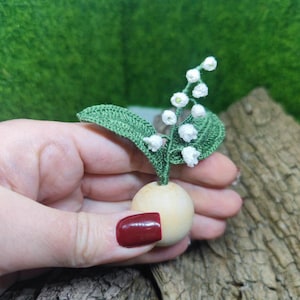 May include: A crocheted lily of the valley flower bouquet with white blossoms and green leaves, attached to a small wooden sphere.