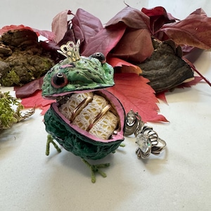 Puede incluir: Un joyero caprichoso en forma de rana verde, coronado con oro. La boca de la rana se abre para revelar un interior forrado de encaje que contiene anillos. Varios anillos de plata con piedras preciosas se colocan cerca, con hojas rojas en el fondo.